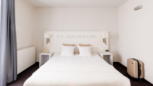 A hotel room with a large bed, two bedside tables, a headboard with the name 'ST.SAUVEUR', and a suitcase on the floor.