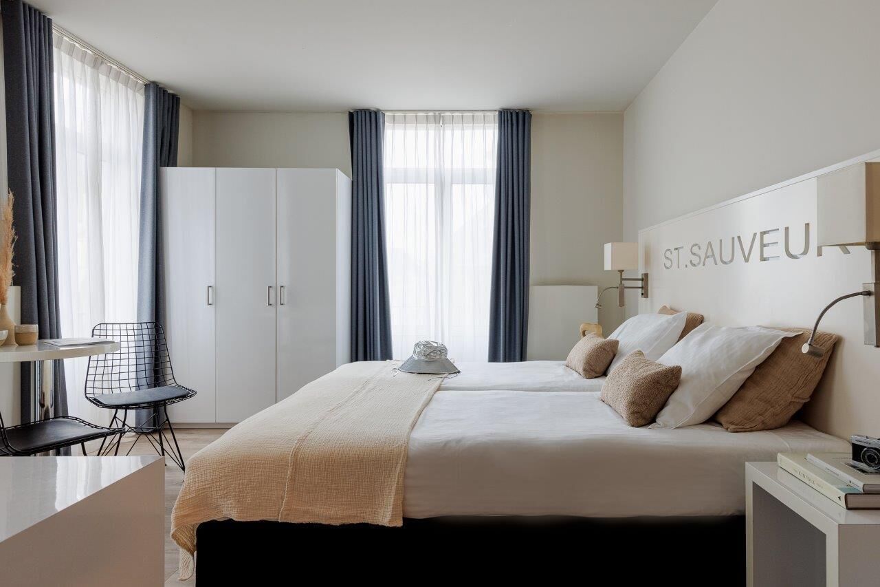Een hotelkamer met een bed, twee nachtkastjes, een bureau met stoel, een garderobe en een raam met gordijnen.