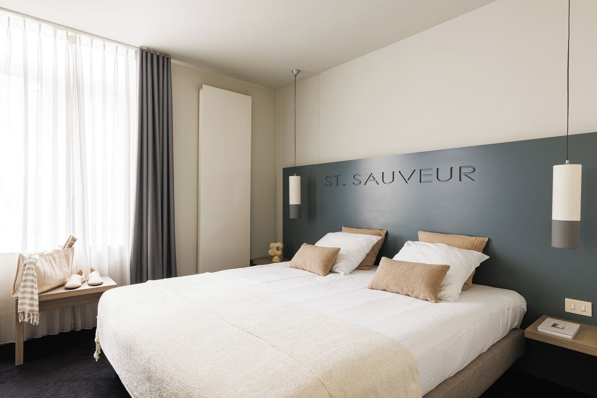A hotel room with a large bed, a bedside table, a chair, and a wall-mounted sign reading 'ST. SAUVEUR'.