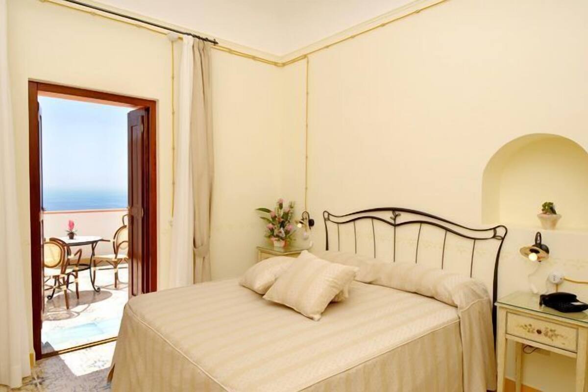 Une chambre d’hôtel avec un lit, une table de chevet, une commode, une petite table avec des chaises et une vue sur la mer par une porte ouverte.