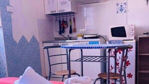 Un lit bien fait, avec des serviettes pliées, une couverture à rayures rouges et blanches, une petite table à manger avec des chaises, un four à micro-ondes et un coin cuisine avec des placards et un pare-feu en carreaux.