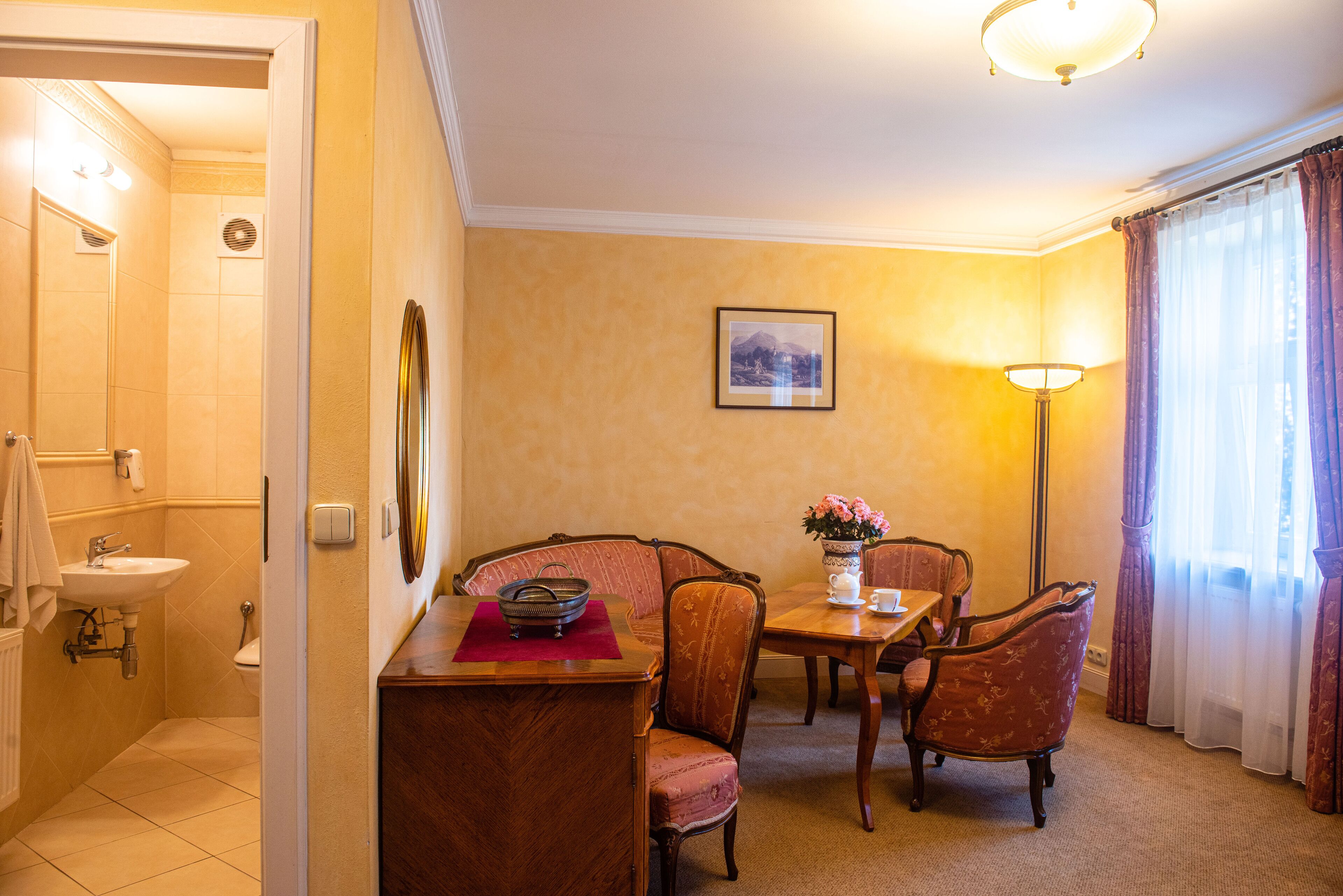 Una habitación de hotel con baño, un comedor con mesa y sillas, y un ventanal con cortinas.