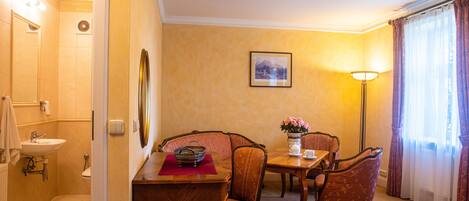 Una habitación de hotel con baño, un comedor con mesa y sillas, y un ventanal con cortinas.