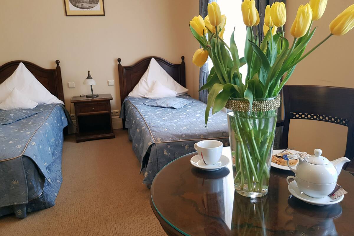 Une chambre d’hôtel avec deux lits, une table ronde avec une théière et une tasse, et un vase rempli de tulipes jaunes.