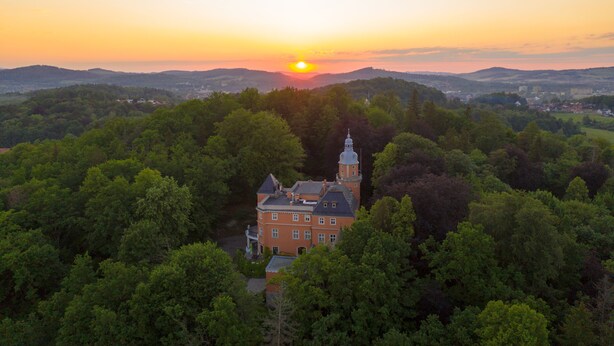 A large, historic building surrounded by dense forest, with a clear view of the sunset in the background. - Palac Paulinum (Jelenia Gora)
