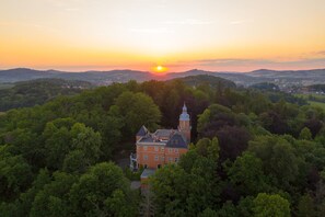 A large, historic building surrounded by dense forest, with a clear view of the sunset in the background. - Palac Paulinum (Jelenia Gora)