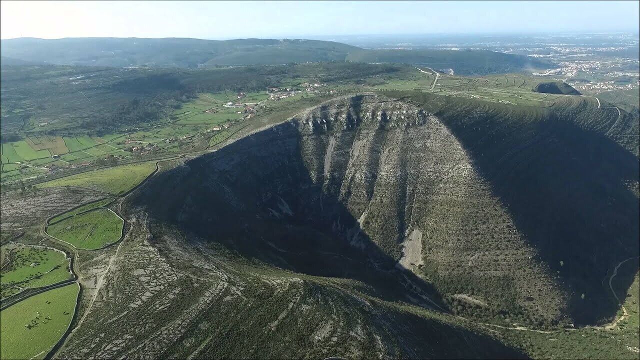An aerial view of a large, steep-sided geological formation surrounded by green fields and a few scattered buildings.