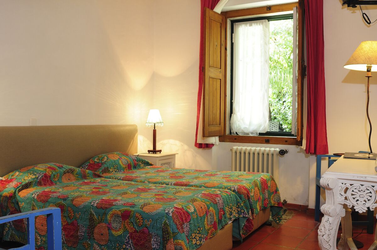 Un dormitorio con dos camas, una ventana con cortinas rojas, una mesita de noche con una lámpara y un radiador.