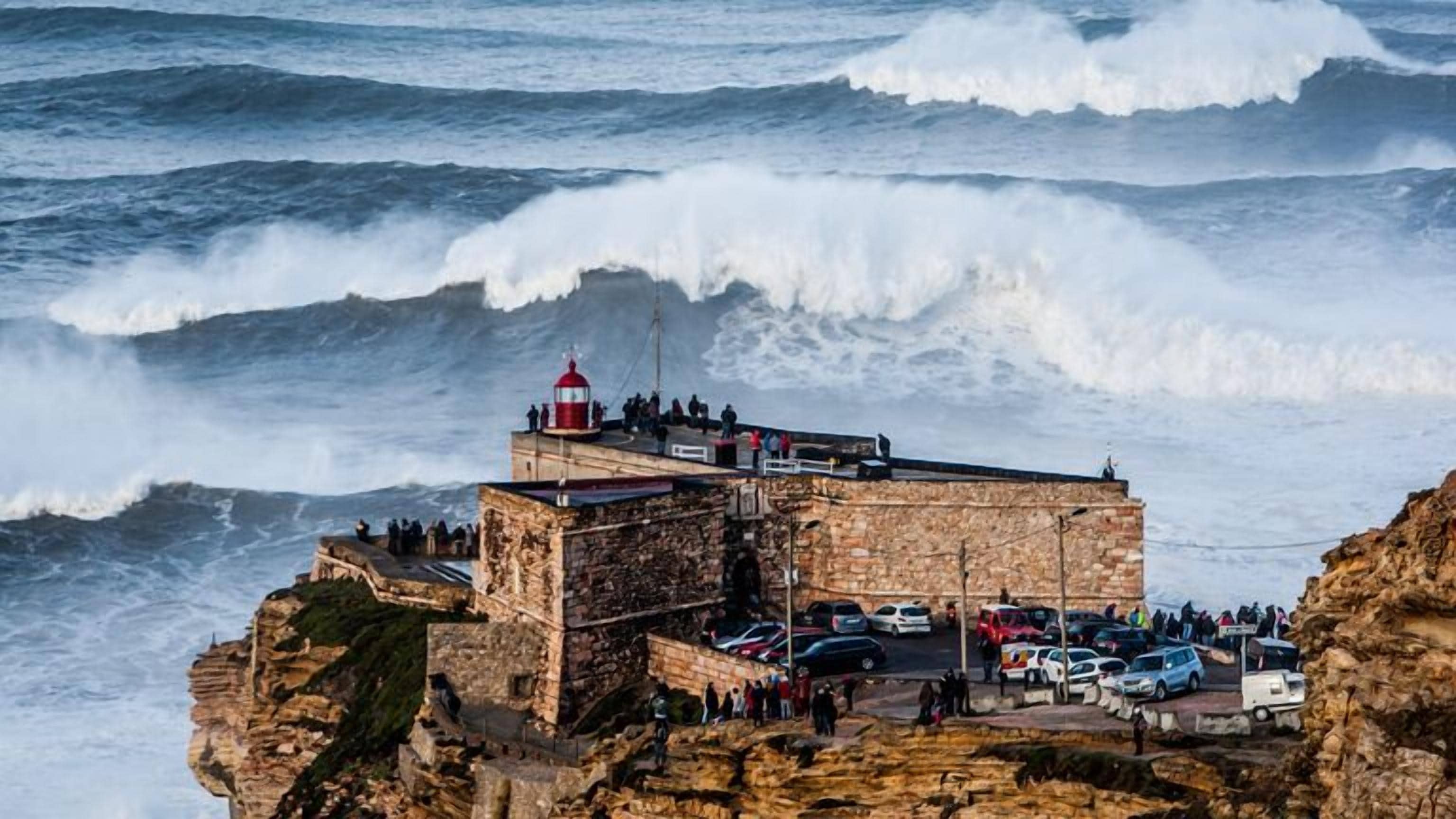 Un faro costero con estructura de piedra, una plataforma de observación y un área de estacionamiento con coches.