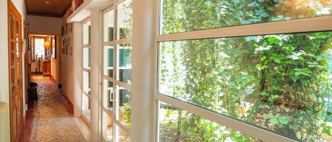 A corridor with a stone floor, large windows with a view of greenery, and a wooden ceiling.