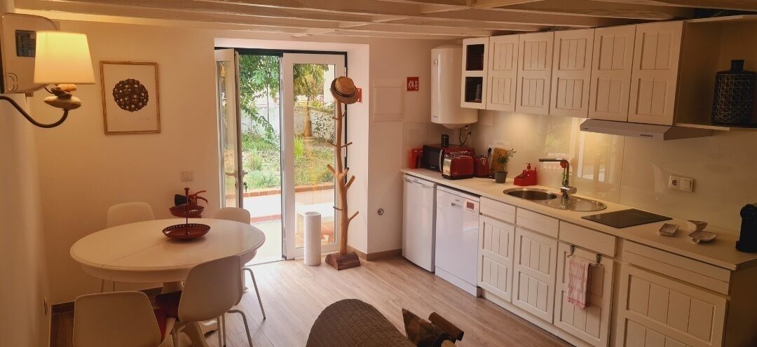 A compact kitchen with white cabinets, a dining table with chairs, and a small countertop with a sink and a kettle.