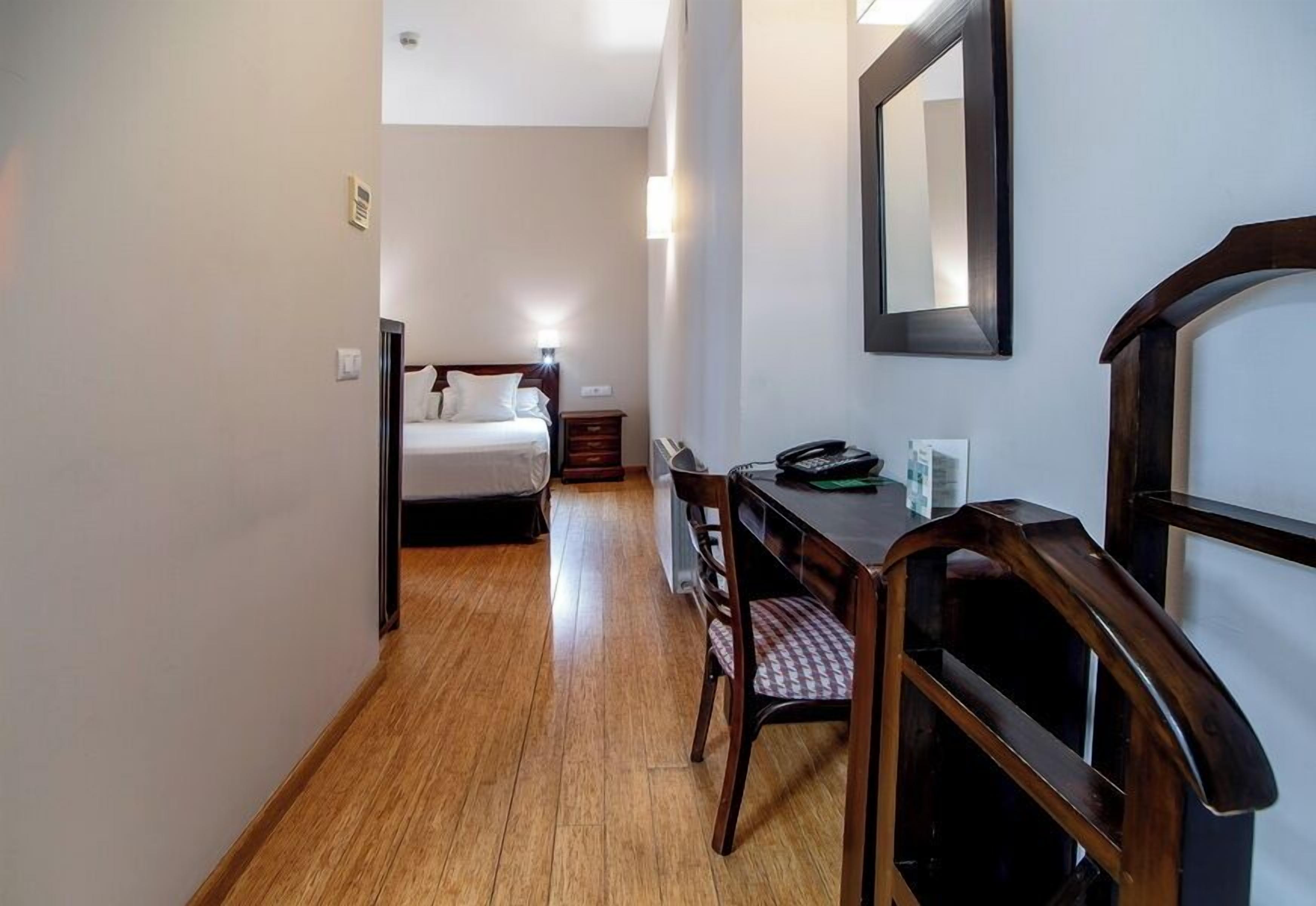 Habitación de hotel con piso de madera, cama con ropa blanca, escritorio con silla, teléfono y espejo en la pared.