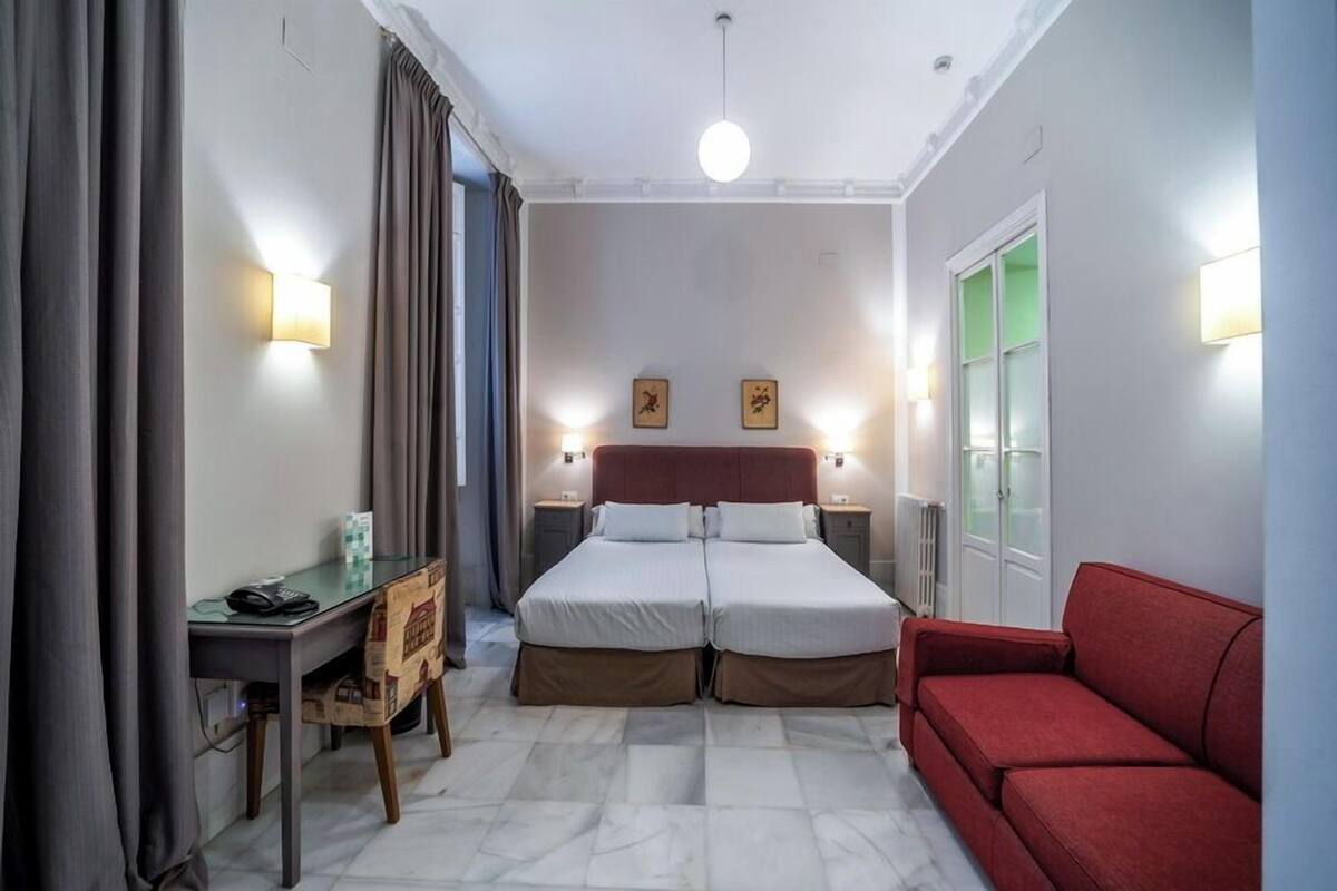 Una habitación de hotel con una cama, un escritorio, un sofá rojo y dos lámparas fijadas a la pared.