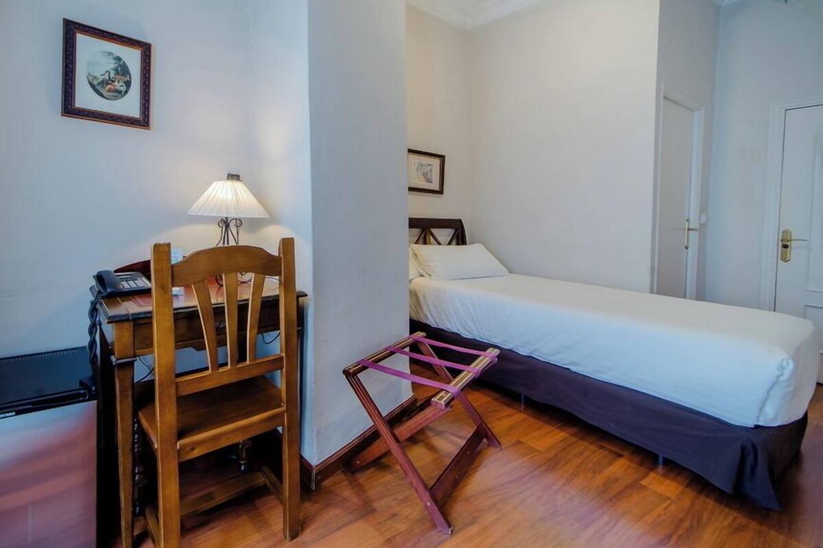 Una habitación pequeña con un escritorio de madera, una silla, una cama y una lámpara de pie.