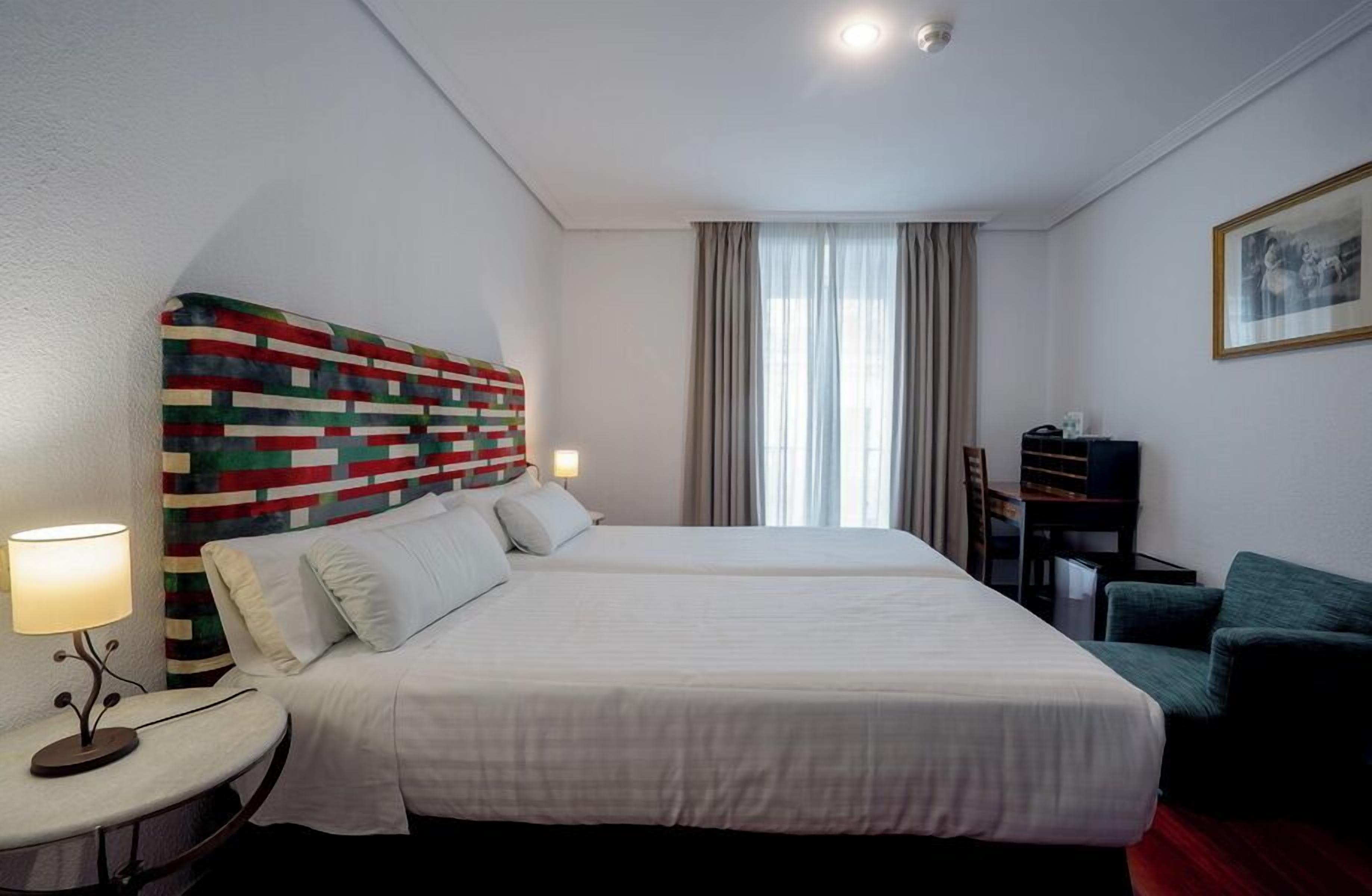 A hotel room with a large bed, a desk, a chair, a lamp, and a colorful headboard.