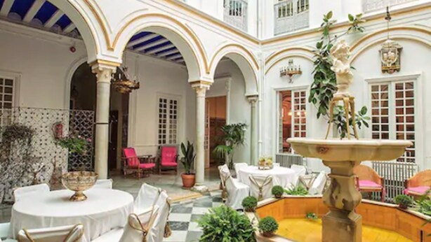 Un patio amplio con paredes blancas, arcos y una fuente central. Mesas y sillas están dispuestas para comer.