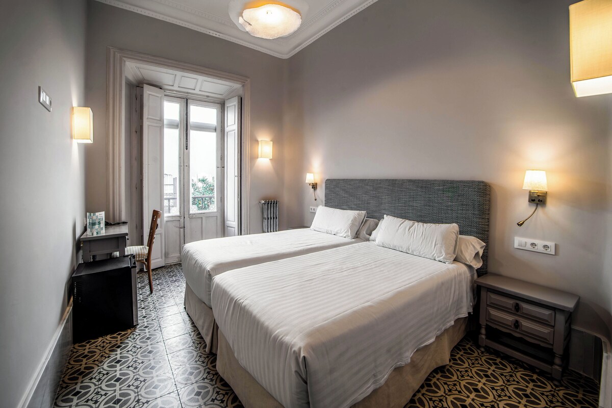 Habitació d'hotel amb dos llits, una tauleta de nit, un armari, una cadira i una finestra amb vistes.