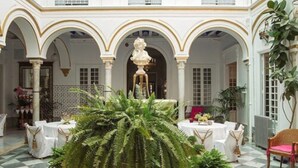 Un patio con columnas blancas, arcos y una fuente central. Se han dispuesto mesas y sillas para comer.