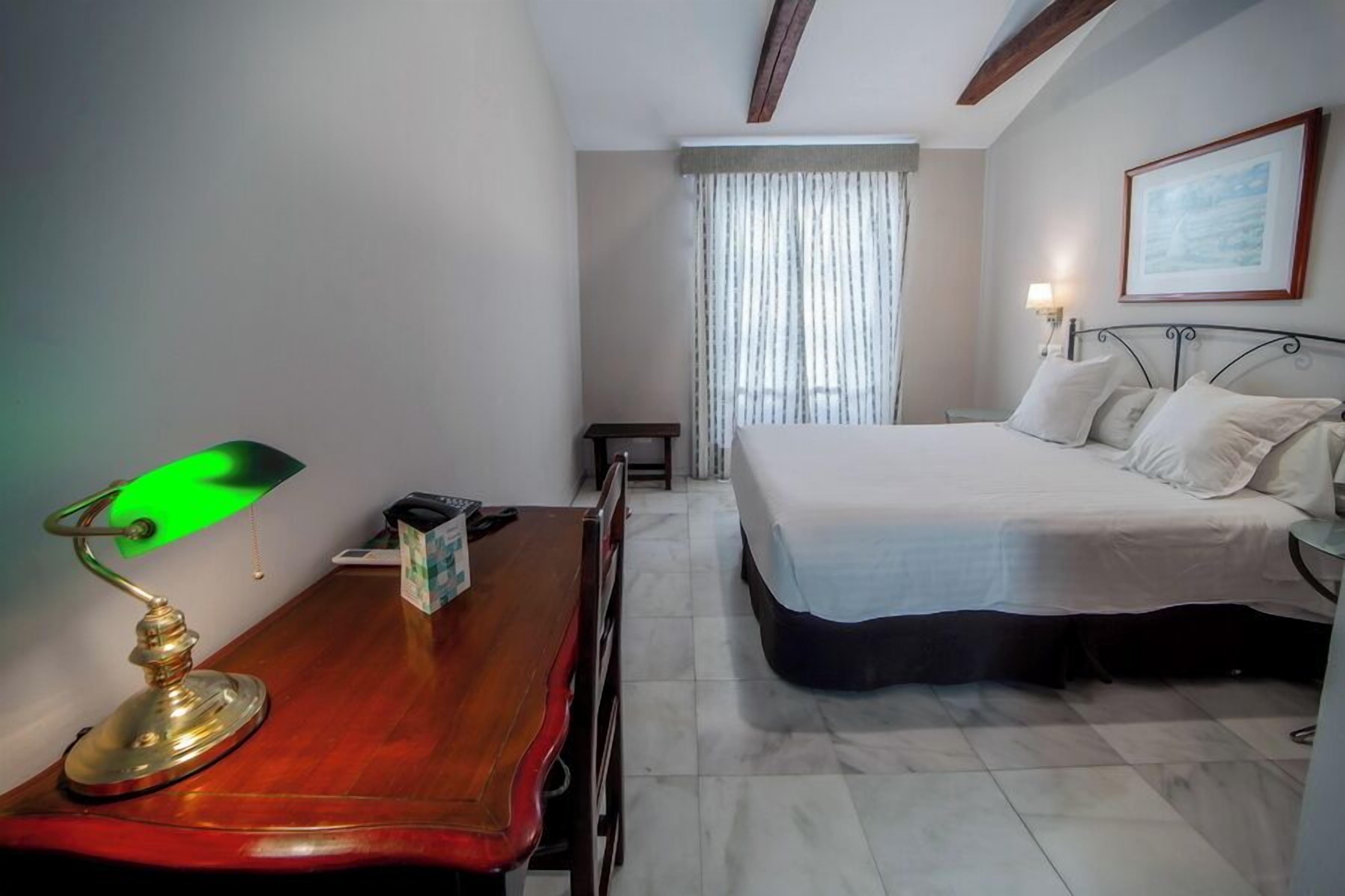 Quarto de hotel com uma cama, uma escrivaninha com abajur, uma mesinha, uma janela com cortinas e um quadro na parede.