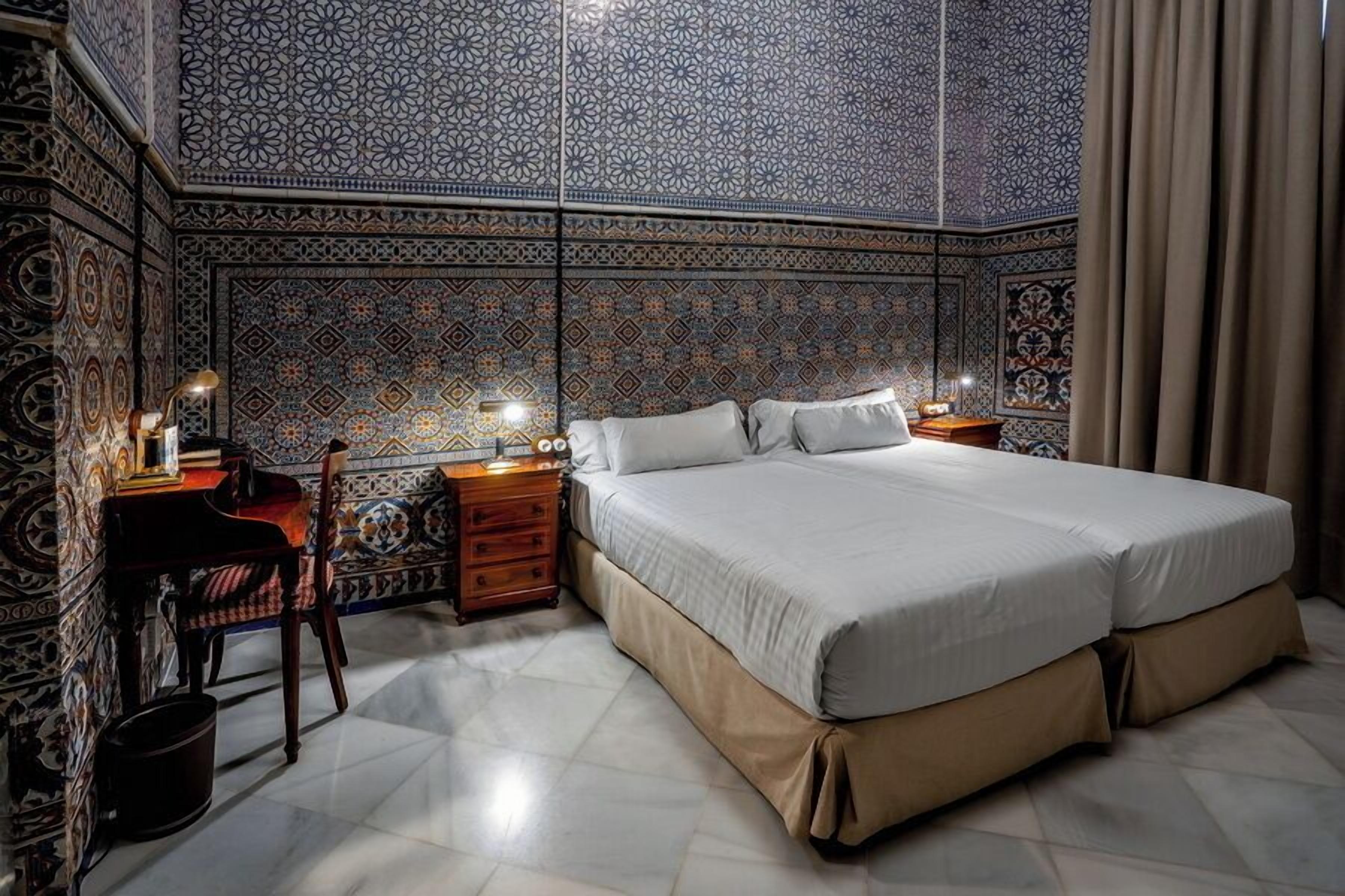 A bedroom with a large bed, a wooden nightstand, a chair, and a patterned wall.