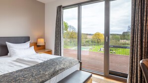 Une chambre d’hôtel avec un grand lit, une table de chevet avec une lampe et une vue sur un balcon doté d’une balustrade et de plantes.