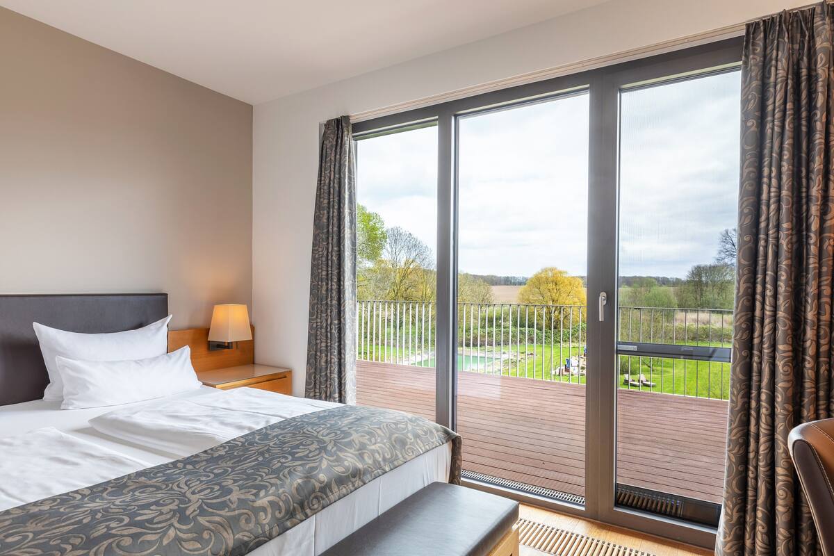 Une chambre d’hôtel avec un grand lit, une table de chevet avec une lampe et une vue sur un balcon doté d’une balustrade et de plantes.