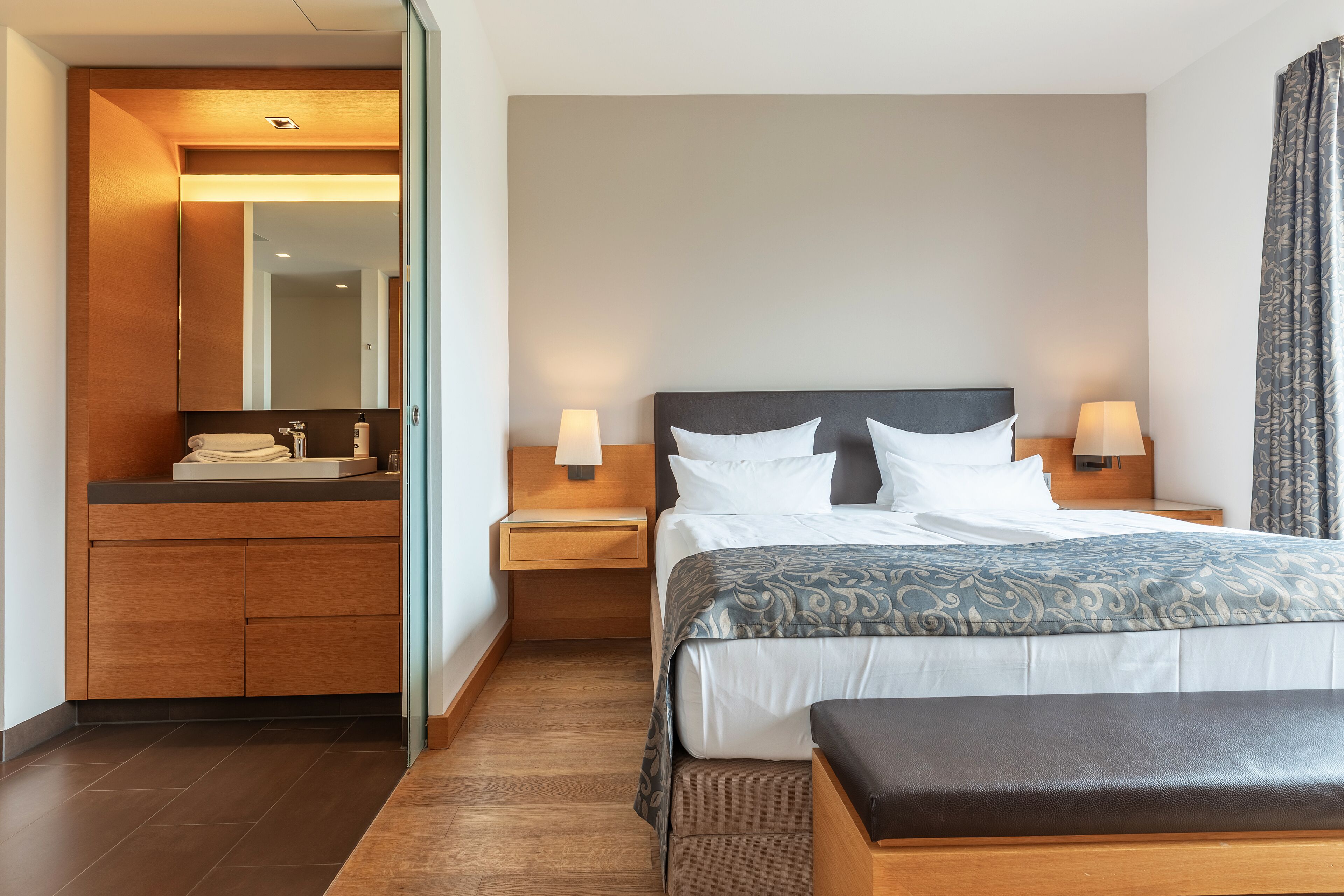 A hotel room with a bed, bedside tables, a mirror, a wooden bench, and a bathroom with a sink and mirror.