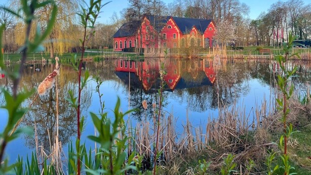 Un bâtiment rouge avec un toit noir, entouré d’arbres et bordé d’un lac paisible.