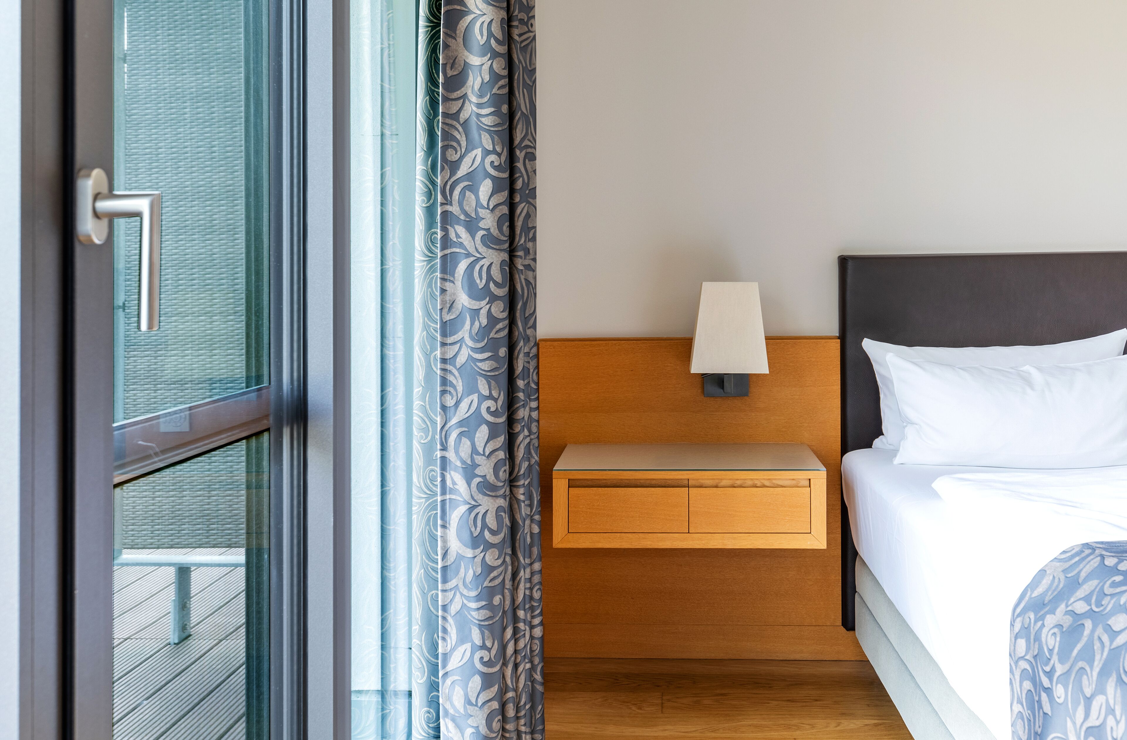 A hotel room with a bed, a nightstand with a lamp, a wooden headboard, and a view through a glass door.