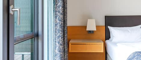 A hotel room with a bed, a nightstand with a lamp, a wooden headboard, and a view through a glass door.