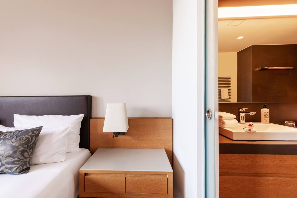 Une chambre d’hôtel comprenant un lit, une table de chevet, une lampe et donnant sur la salle de bain, où l’on aperçoit un lavabo et un miroir.