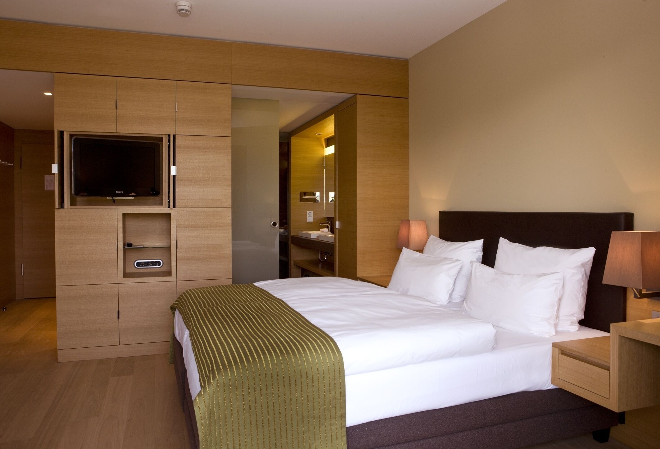 A hotel room with a bed, a TV mounted on a wooden cabinet, a bedside table with a lamp, and a mirror on the wall.