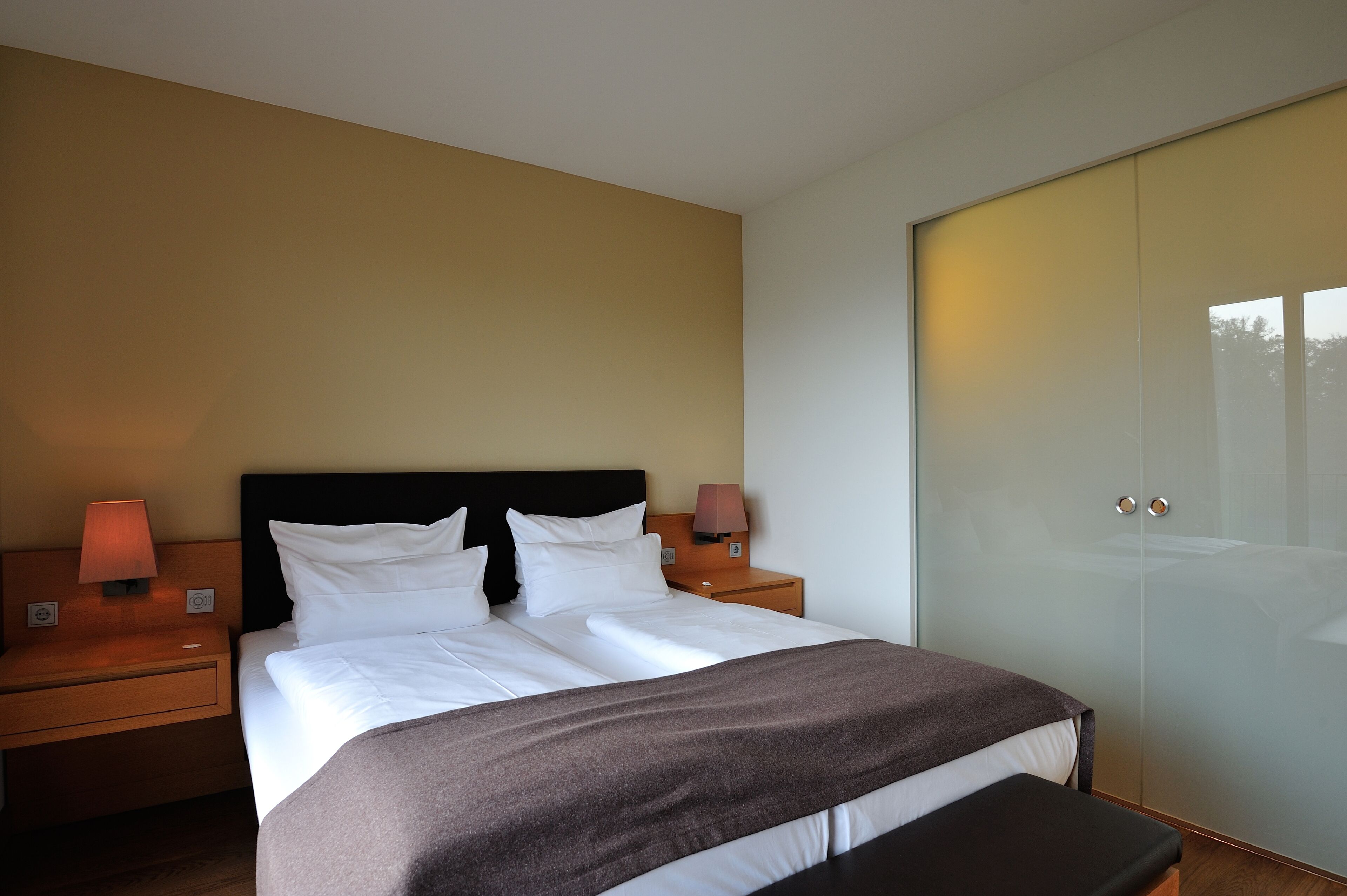 A modern hotel room with a large bed, two bedside lamps, a nightstand, and a wardrobe.