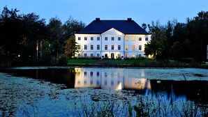 Un grand bâtiment blanc, surmonté d’un toit sombre, entouré d’arbres et bordé par une étendue d’eau dans laquelle se reflète sa silhouette.