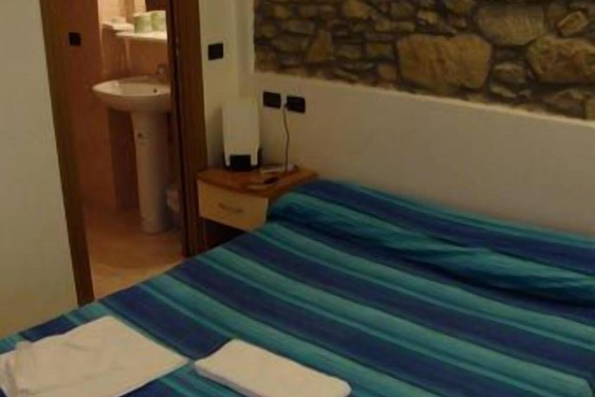 Un dormitori amb un llit, una tauleta de nit, una paret de pedra i un bany visible a través d'una porta oberta.