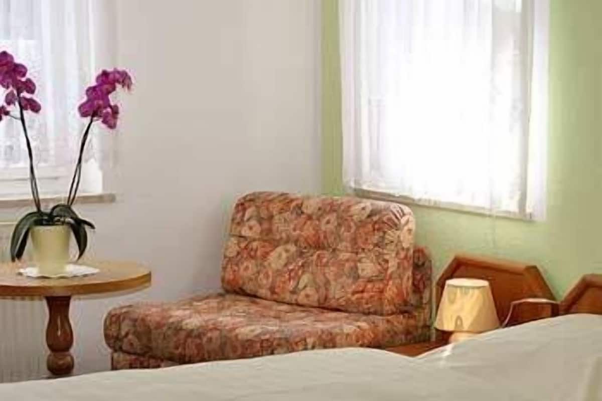 A room with a bed, a floral-patterned chair, a small round table with a potted plant, and a lamp.