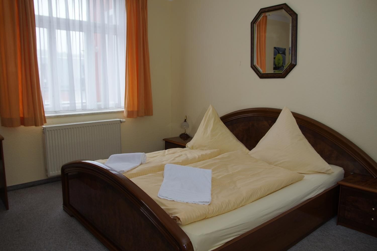 Un dormitorio con una cama de madera, dos almohadas, una mesita de noche, un espejo y una ventana con cortinas.