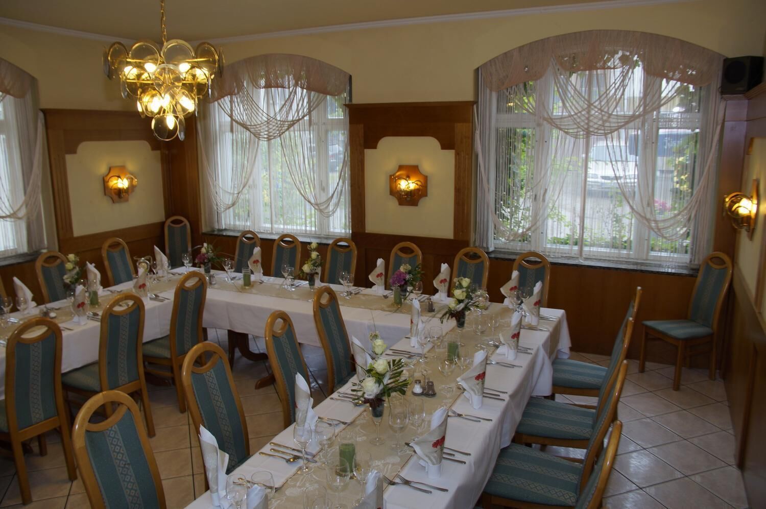 Un comedor con mesas largas dispuestas para varios comensales, sillas con asientos acolchados y apliques decorativos en la pared.