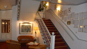 A hotel lobby with a staircase, a sofa, a lamp, and framed certificates on the wall.