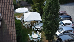 An outdoor seating area with white tables and chairs, a large white tent, and a row of parked cars.