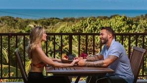 A couple sitting at a wooden table with a basket of fruit and flowers, overlooking a lush garden and the ocean.