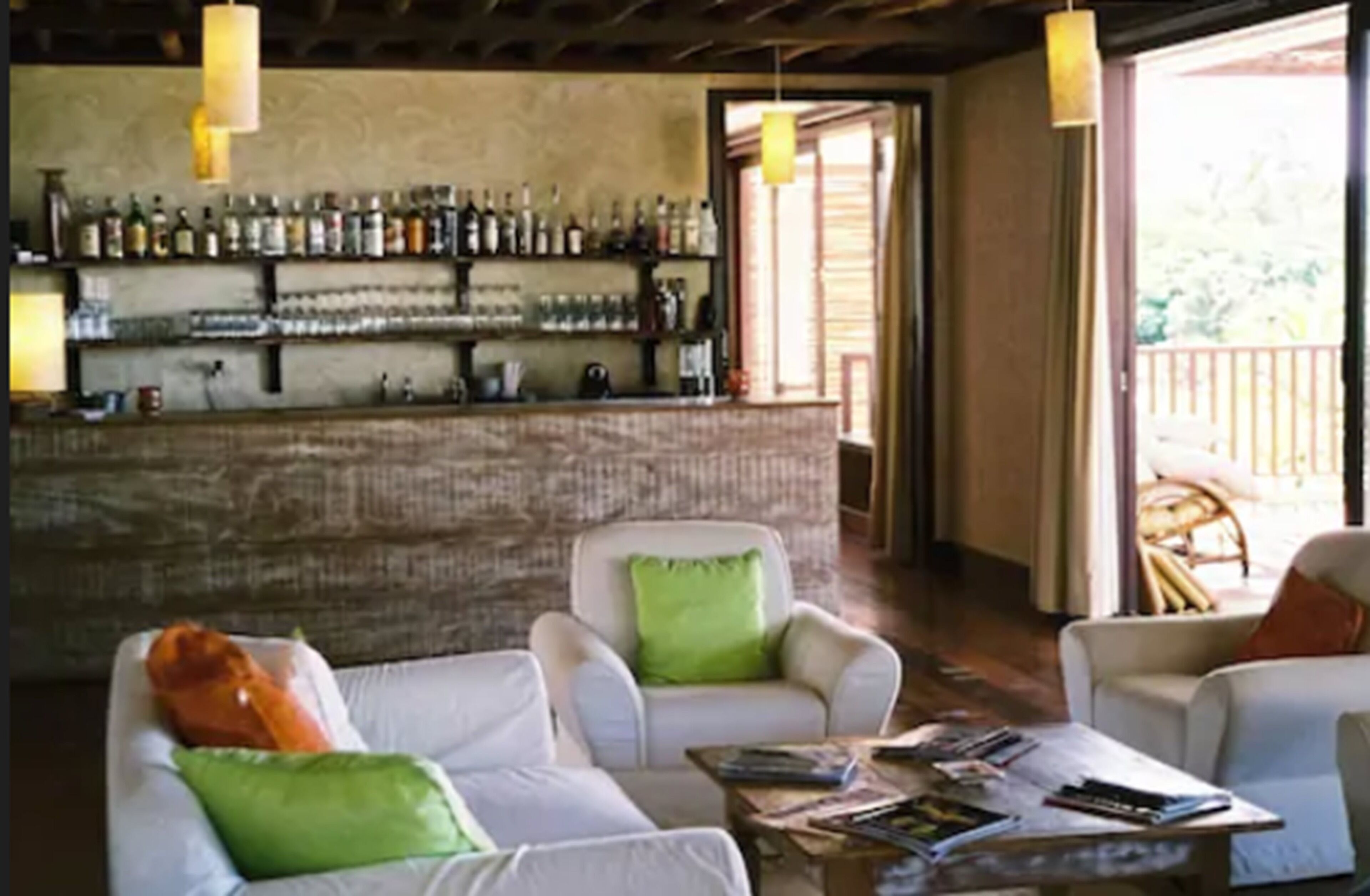 A bar area with shelves of bottles and glasses, a counter, and a seating area with white sofas and green cushions.