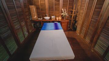 A room with a bed, wooden shutters, a small table with a bowl, candles, and a vase with flowers.