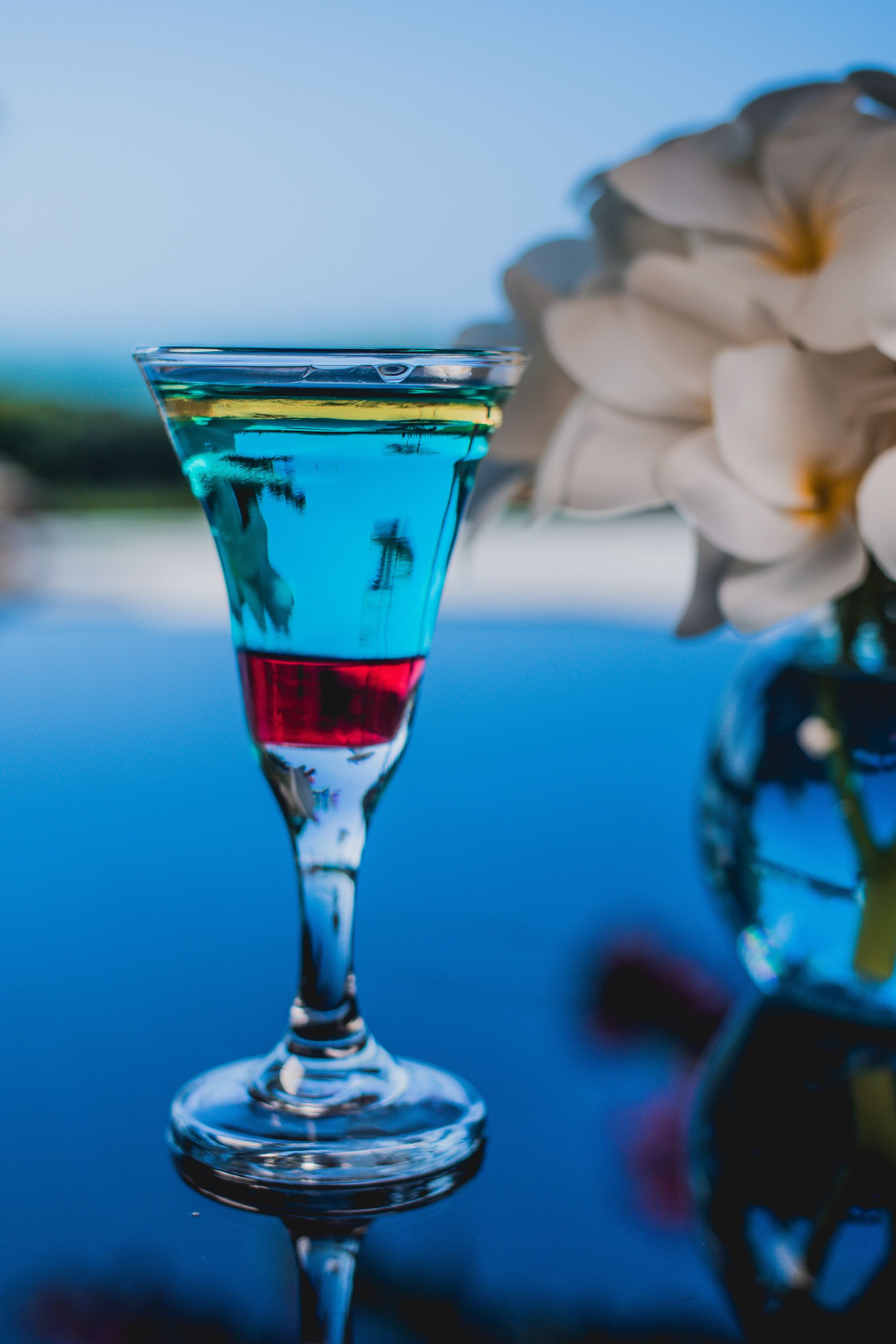 A clear glass with a blue liquid, a white flower, and a blurred background.