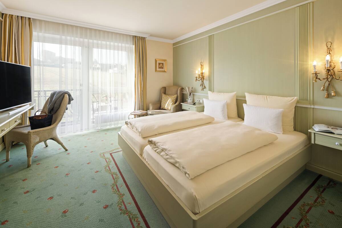 A hotel room with a large bed, a desk with a TV, a chair, a nightstand, and a window with curtains.