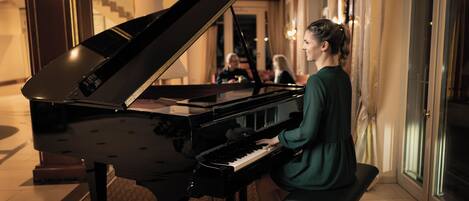 A grand piano in a well-lit room with a bench, a staircase in the background, and a large window.