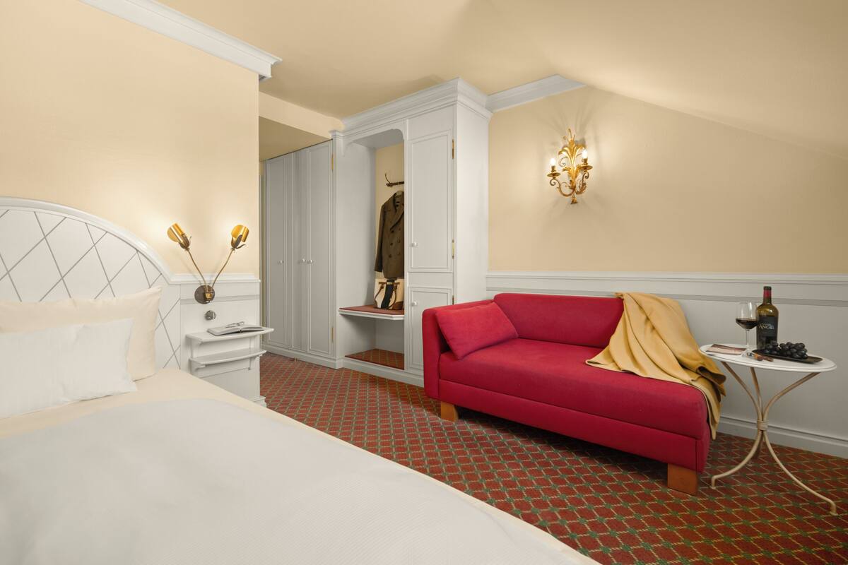 A hotel room with a bed, a red sofa, a small table with a bottle and glasses, and a wardrobe.