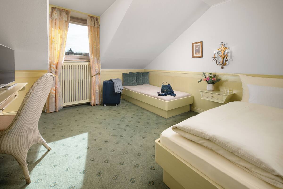 A hotel room with a bed, a desk, a chair, a small sofa, a radiator, a window with curtains, and a wall-mounted light fixture.