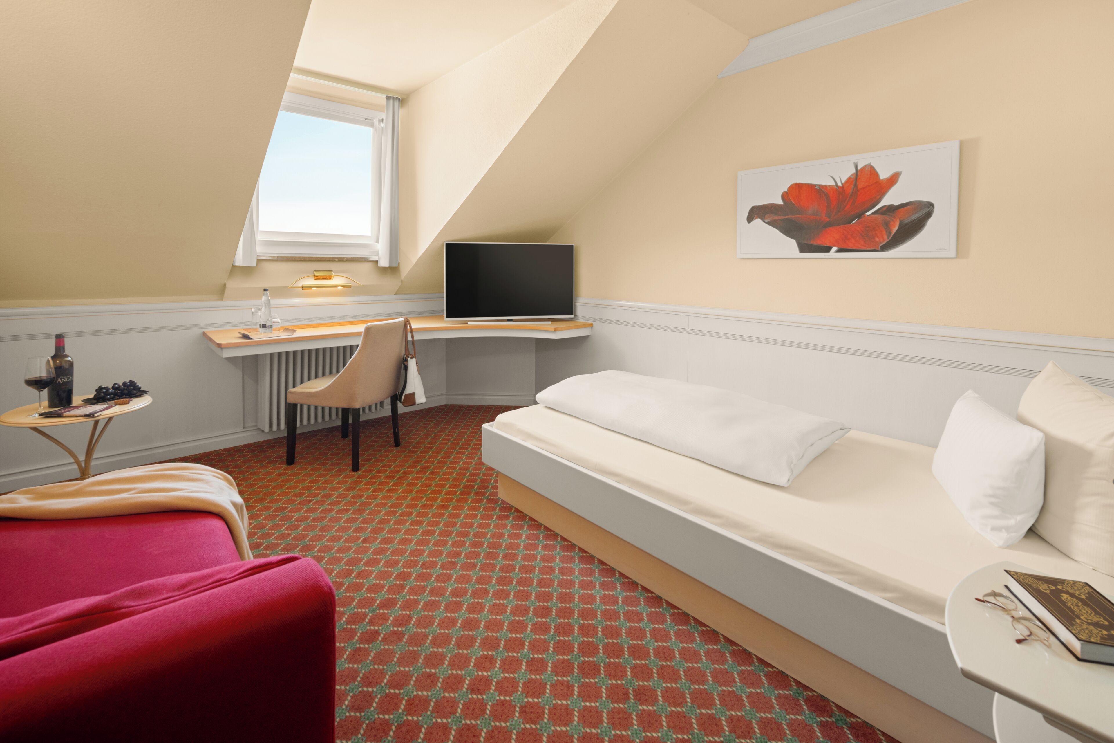 Habitación de hotel con cama, escritorio con televisor, silla, mesita con una botella y ventana.
