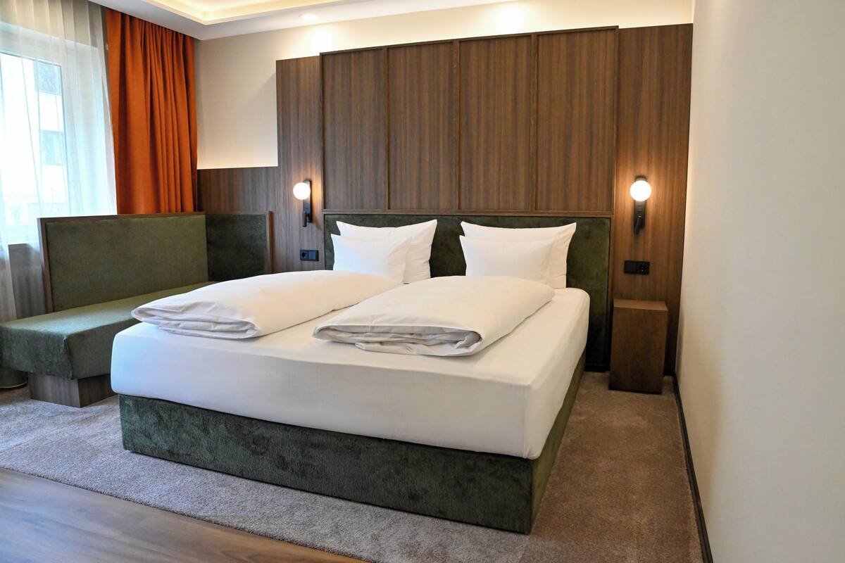 A modern hotel room with a large bed, two bedside tables, a bench, and a view of the outdoors.